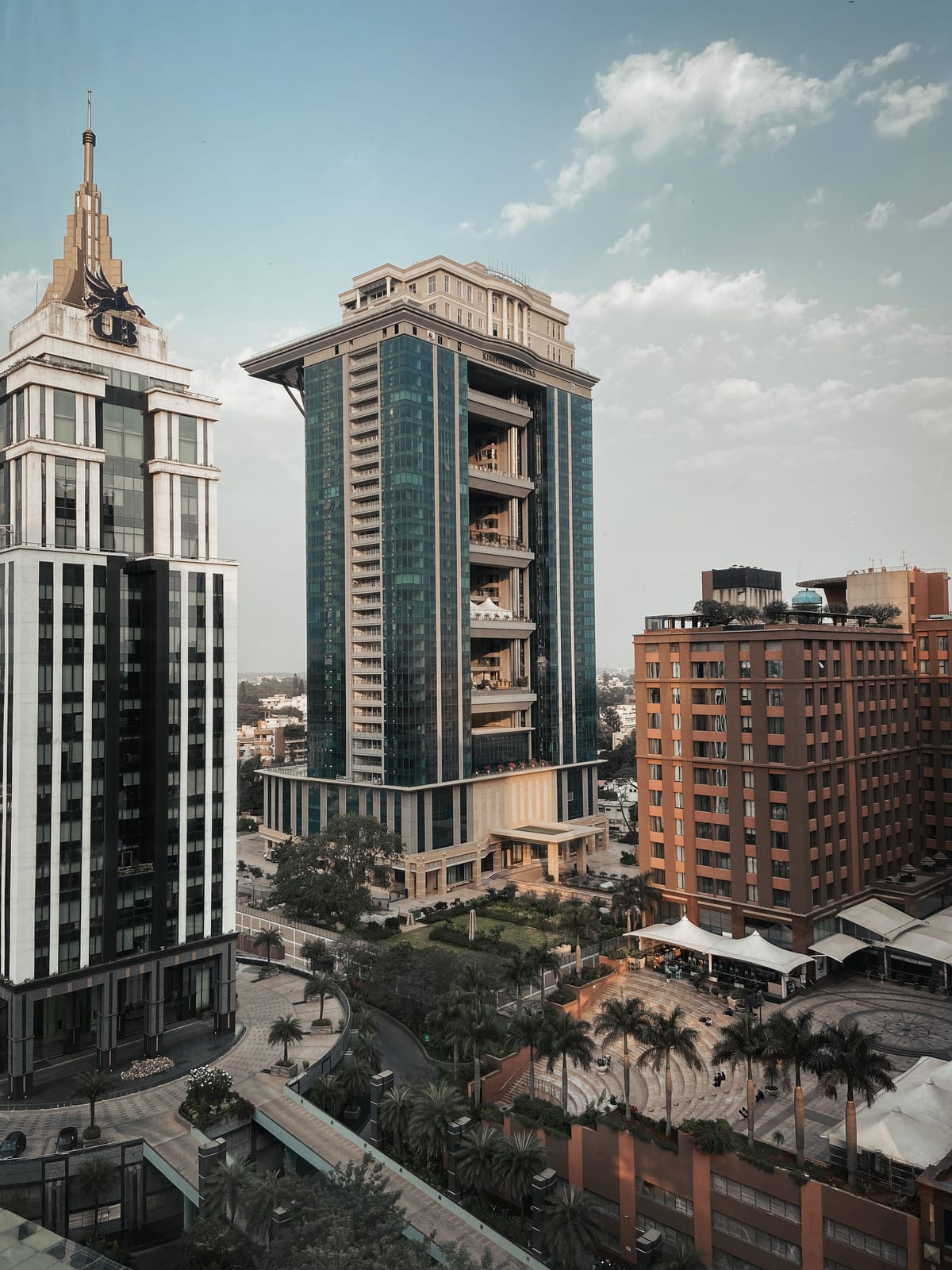 Elevated view across a city business district: a glass-clad tower with stacked balcony terraces stands between a pale stone high-rise and a red-brick block, with a palm-lined plaza and walkways below.